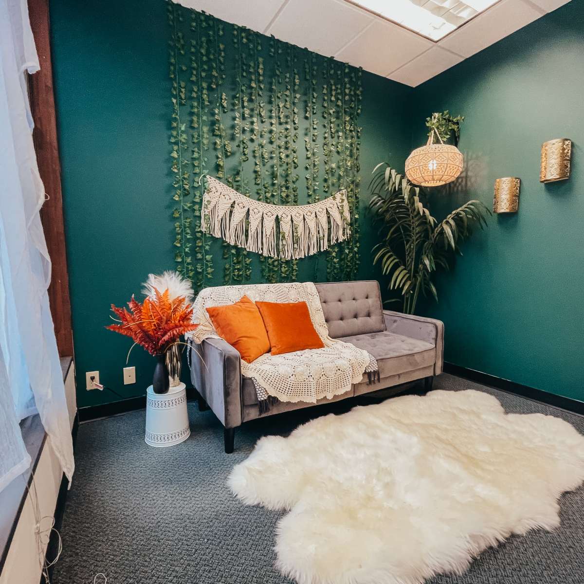 A cozy room featuring a gray couch adorned with orange pillows, a decorative wall hanging, and greenery, alongside a white faux fur rug on a carpeted floor.