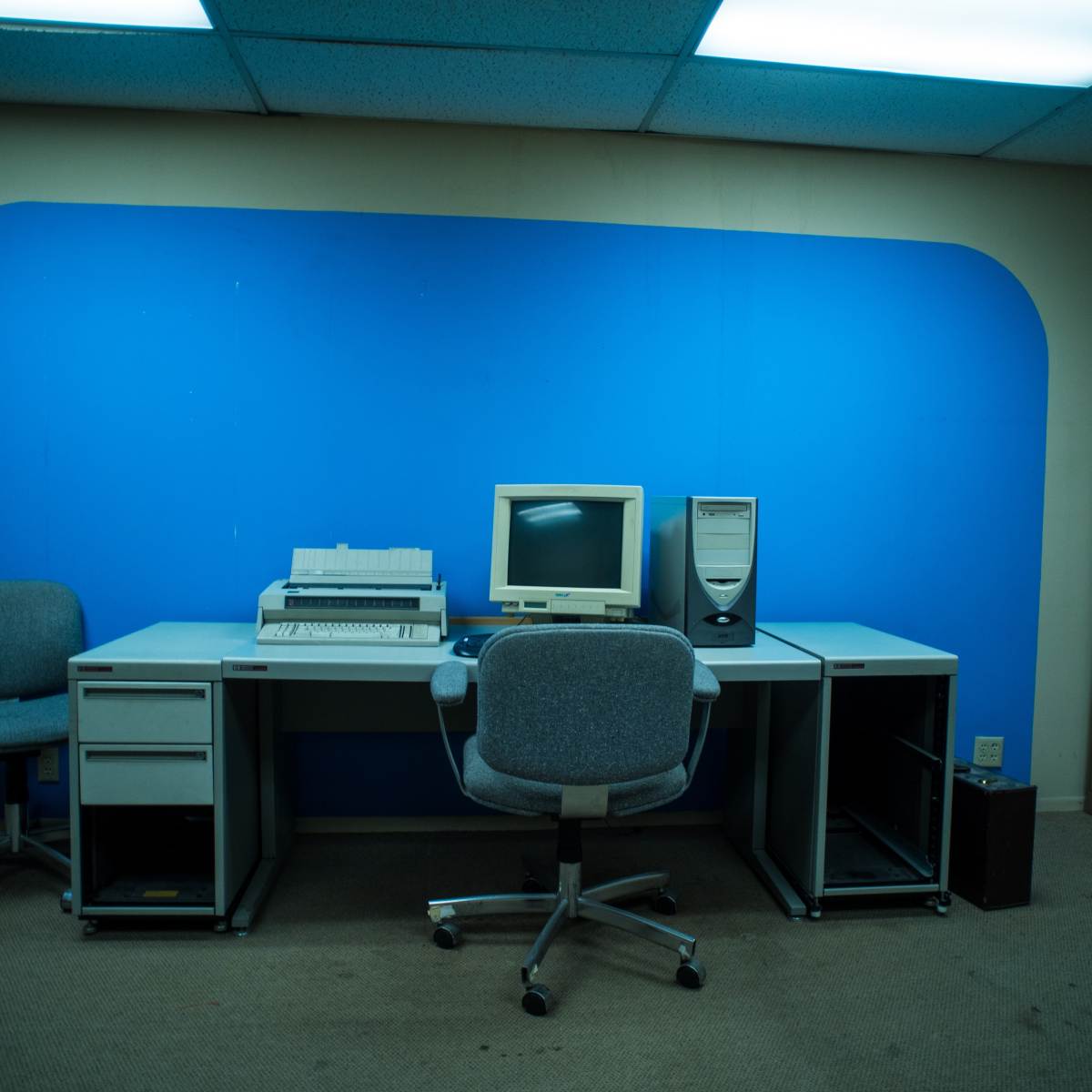 An empty office space features a desk with a computer monitor, a printer, and two chairs against a blue wall.
