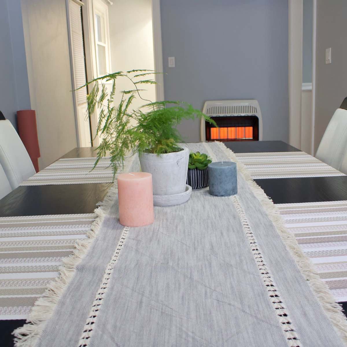A dining table is set with a table runner, candles, a potted plant, and a small decorative bowl, with a heater visible in the background.