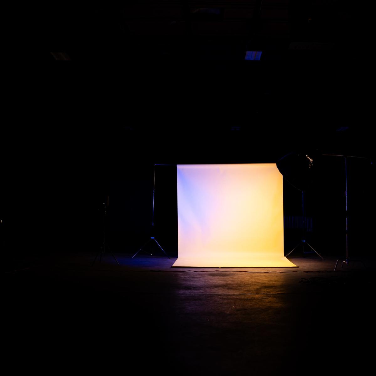 A dimly lit studio features a brightly illuminated backdrop with lighting equipment positioned nearby.