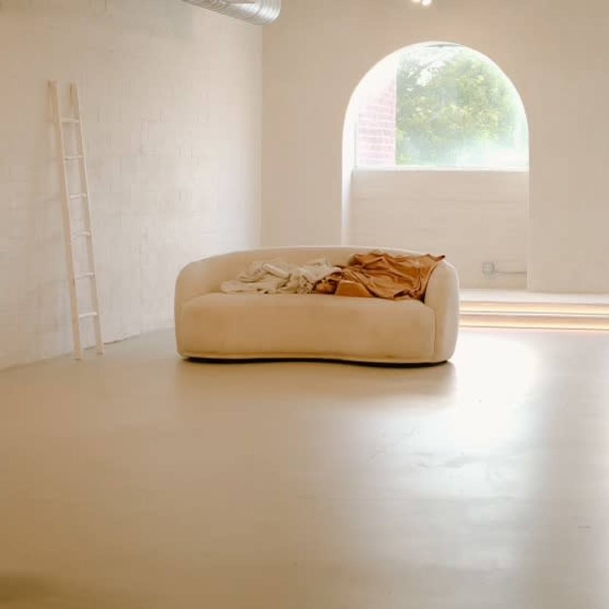 A spacious room features a curved beige sofa with blankets, large arched windows, and a circular platform on the floor.
