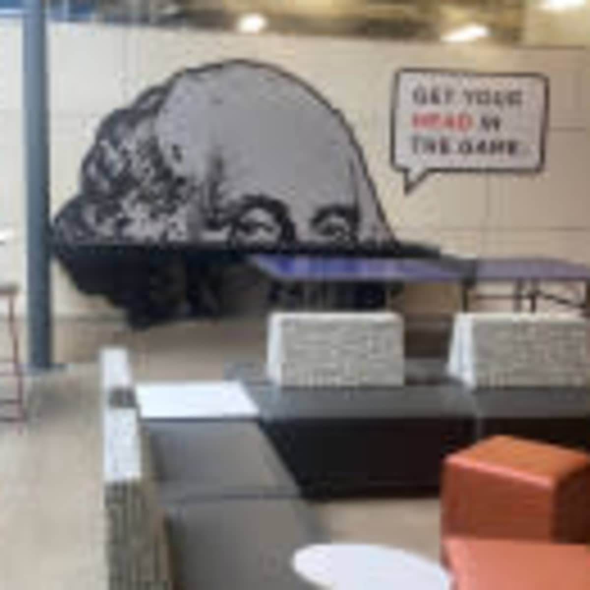 A modern workspace with a large mural of a man's head and a motivational phrase on the wall, along with various seating areas and tables.