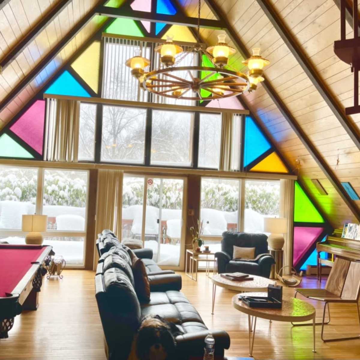 A spacious A-frame living room with colorful stained glass windows, a pool table, and large windows overlooking a snowy landscape.