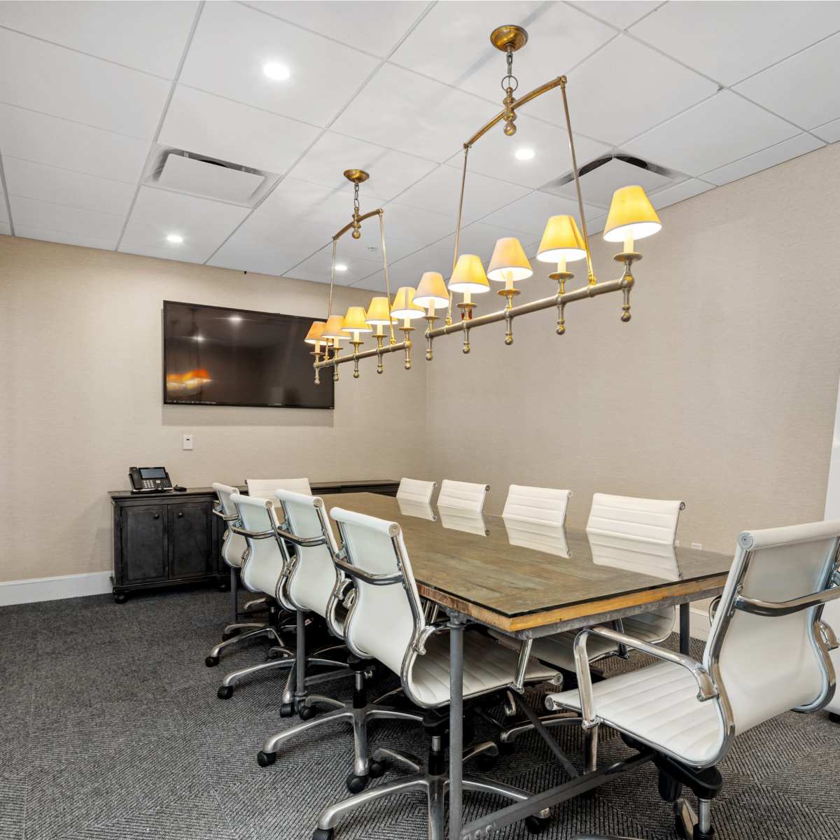 A conference room features a long wooden table surrounded by white chairs, with a row of pendant lights above and a wall-mounted screen on one side.