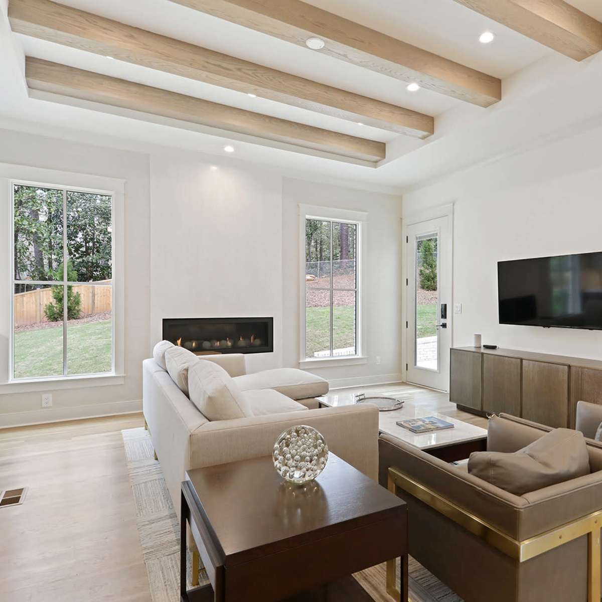 A modern living room with a sectional sofa, television mounted on the wall, and large windows allowing natural light.