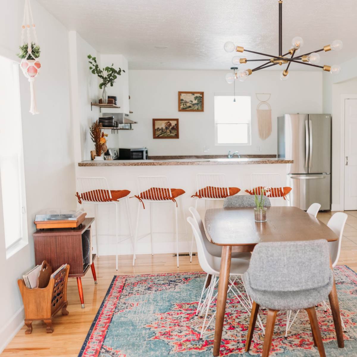 A modern dining area with a wooden table and chairs, set against a backdrop of a light-colored kitchen featuring bar stools and a fridge.