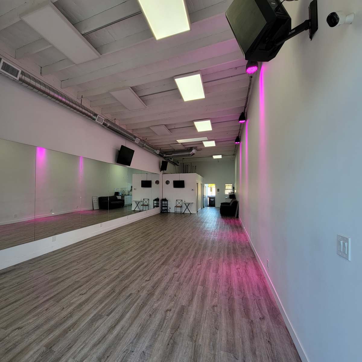 A spacious, empty studio with light wood flooring, white walls, and pink LED lighting along the mirrors.