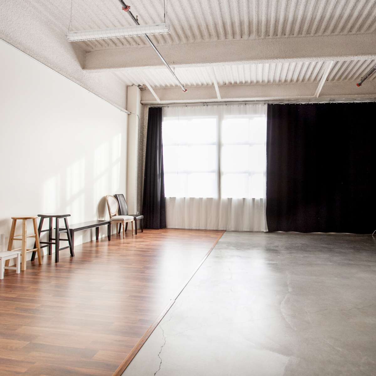 An empty studio space with a wooden floor on one side and a concrete floor on the other, featuring a large window and black curtains.