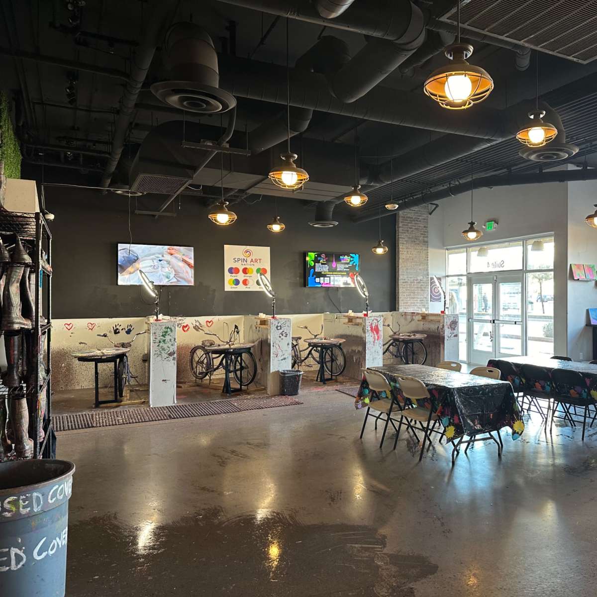 An indoor art studio with tables covered in paint splatters, several easels set up for painting, and light fixtures hanging from the ceiling.
