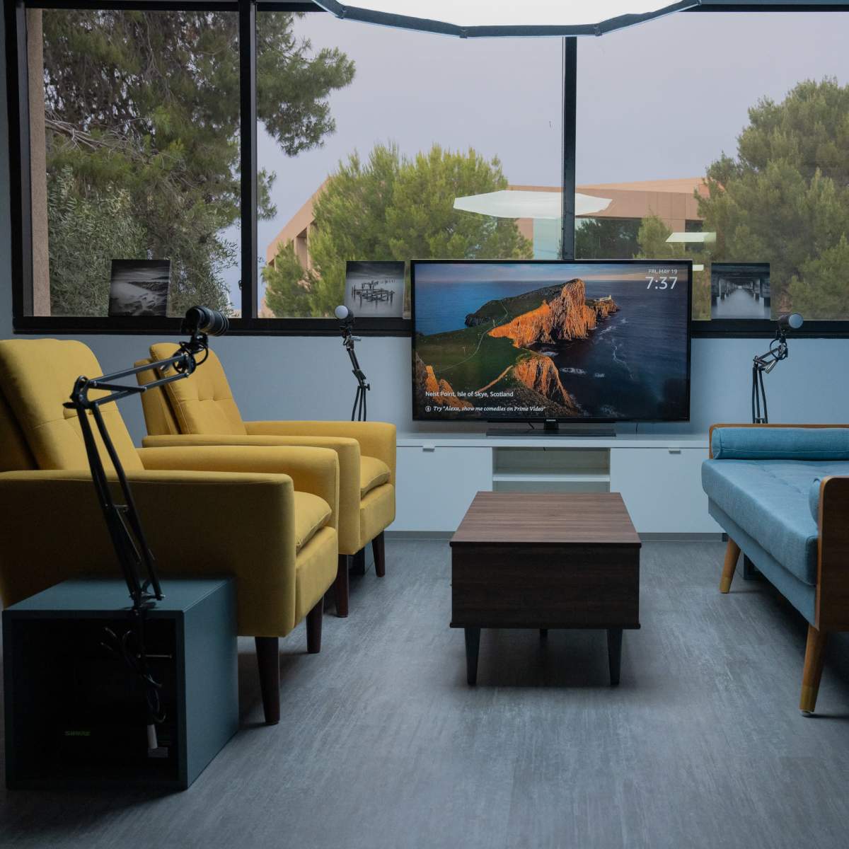 A modern living space features two armchairs, a blue couch, a coffee table, and a television mounted on the wall, all set against a large window with trees outside.