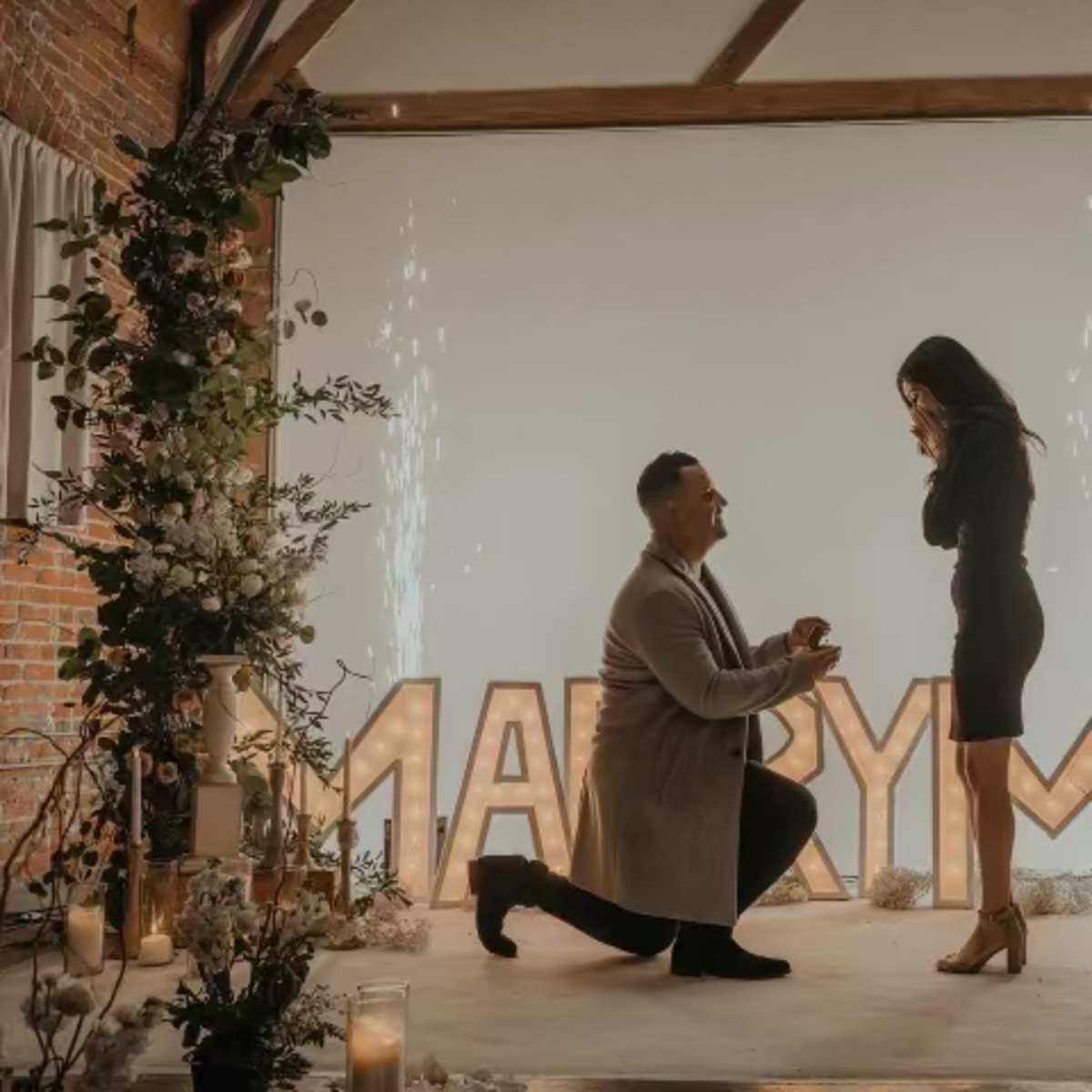 A man is kneeling on one knee proposing to a woman in front of a large "MARRY ME" sign, surrounded by floral decorations and candles.