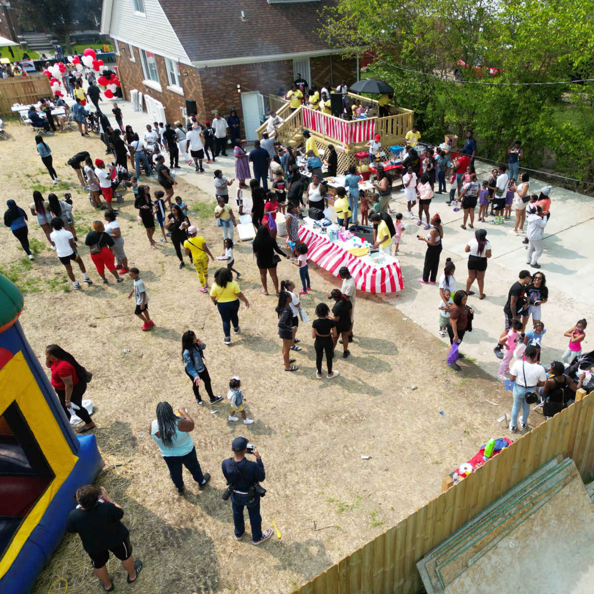 A large group of people gathers in a backyard with a bounce house, tables with food, and festive decorations.