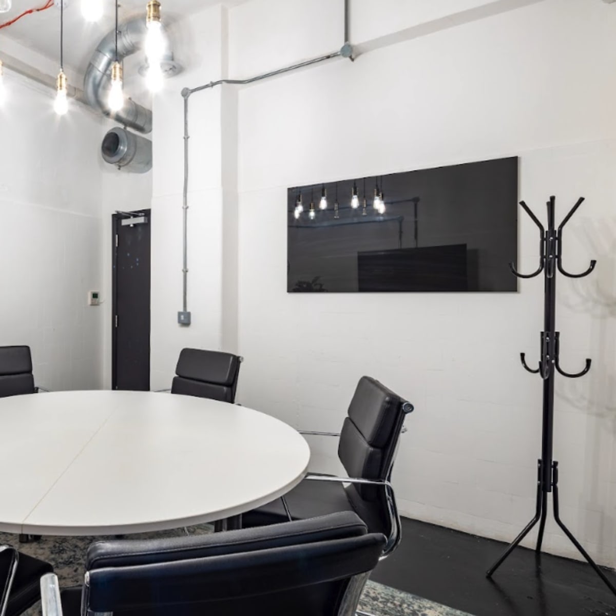 A round white table surrounded by black chairs in a minimalist meeting room with exposed pipes and overhead lights.