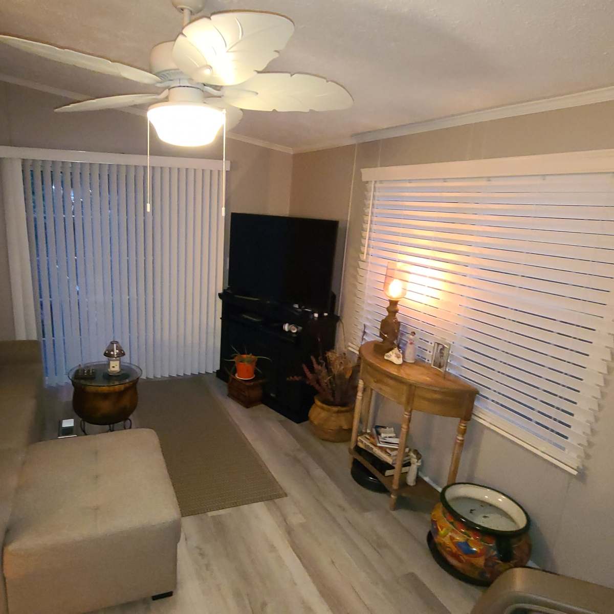 The living room features a sectional sofa with decorative pillows, a television on a black stand, and large windows covered with blinds, alongside a coffee table and a decorative bowl.
