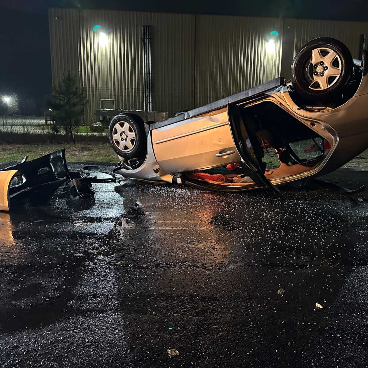 A silver car is overturned on its roof on a dark, wet roadway, with debris scattered nearby.