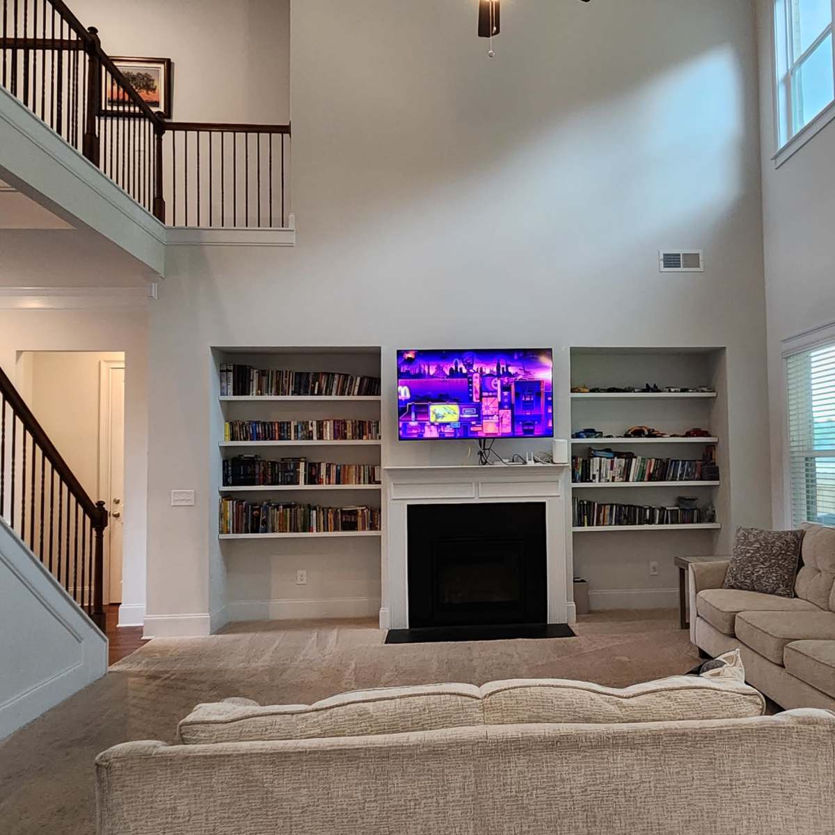 The living room features high ceilings, a fireplace, a large TV mounted on the wall, and bookshelves filled with books.