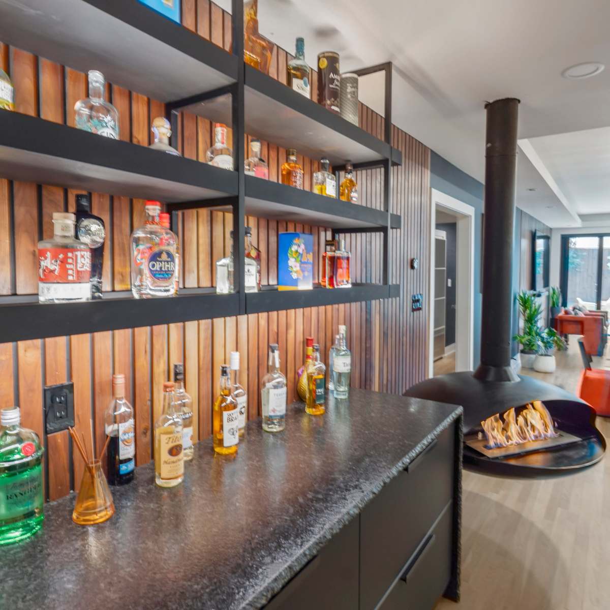 A modern home bar with a black countertop, a selection of various liquor bottles neatly arranged on open shelves, and a round fire pit in the foreground.
