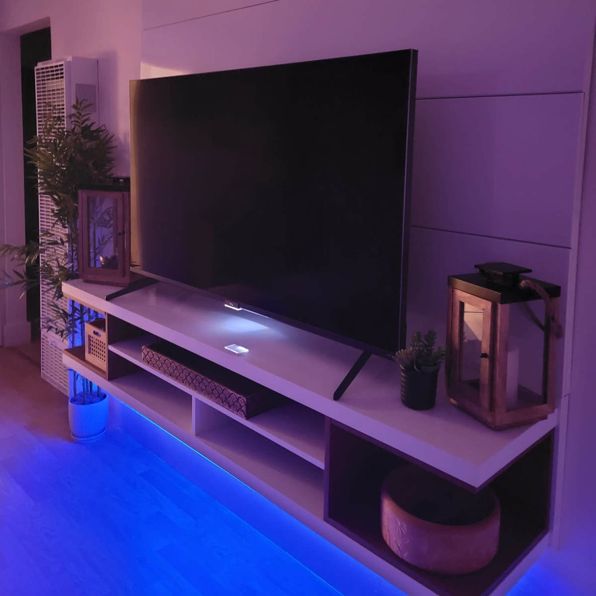 A large TV is mounted on a wall-mounted shelf above a sleek, modern entertainment unit with ambient lighting.