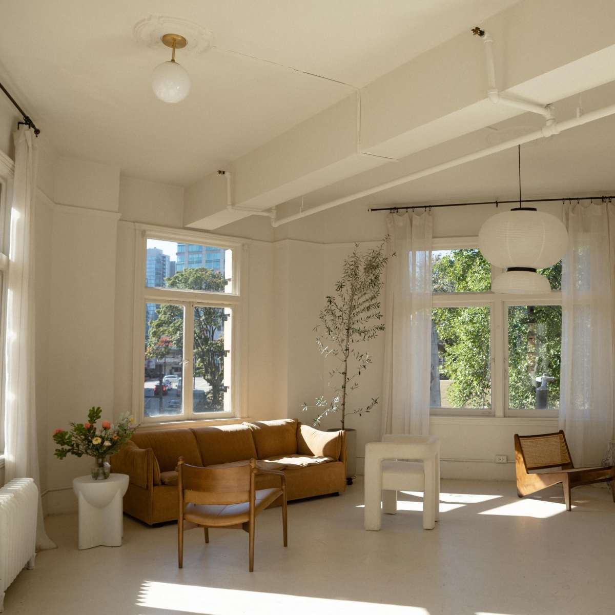 A bright, minimalist living space with large windows, a beige couch, a wooden chair, a small table with a vase, and light-colored walls.