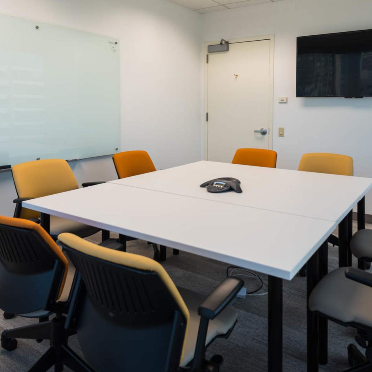 A conference room features a large white table surrounded by six chairs, a wall-mounted television, and a glass whiteboard.