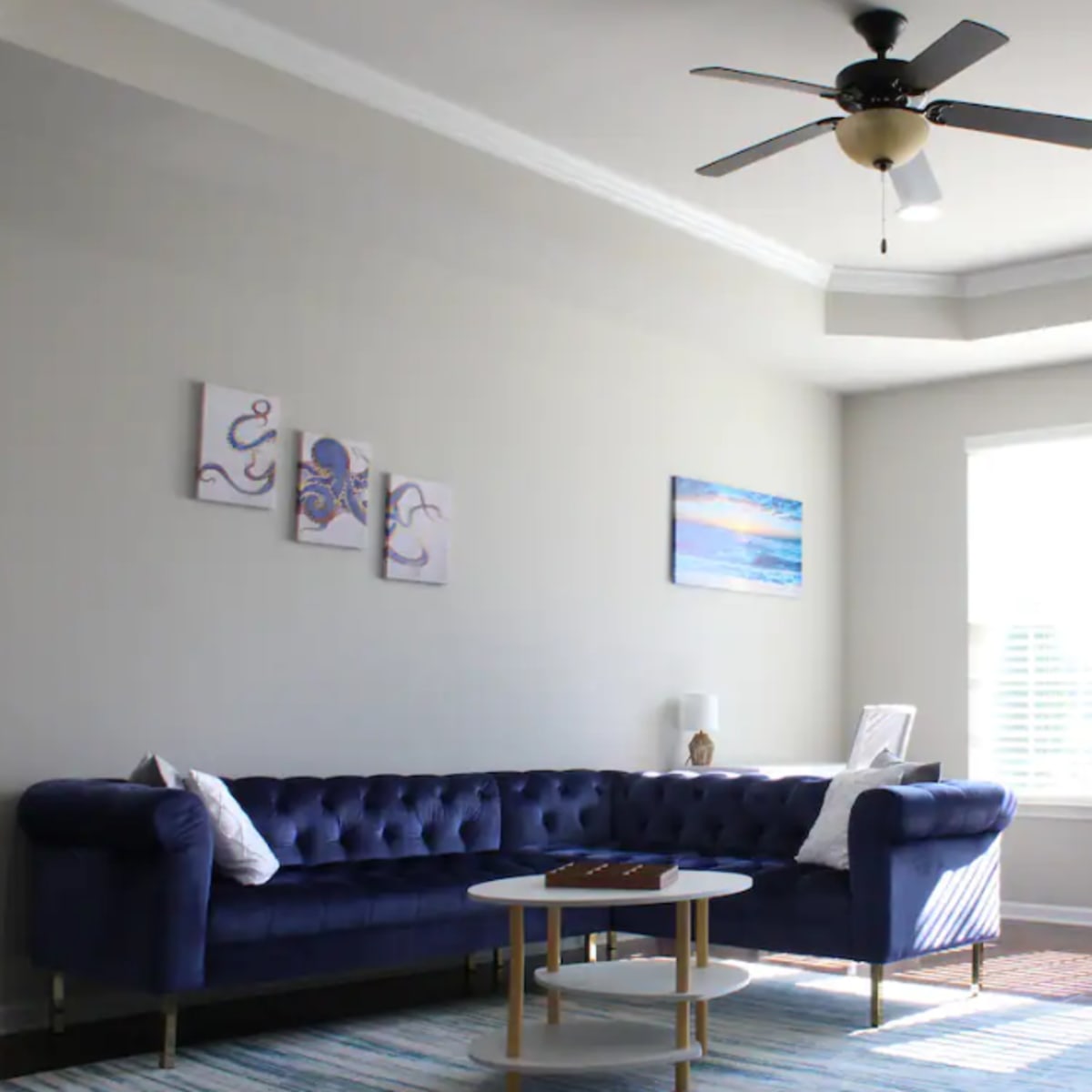 A blue tufted sofa is positioned in a well-lit living room with large windows and a ceiling fan, accompanied by a coffee table and wall art.