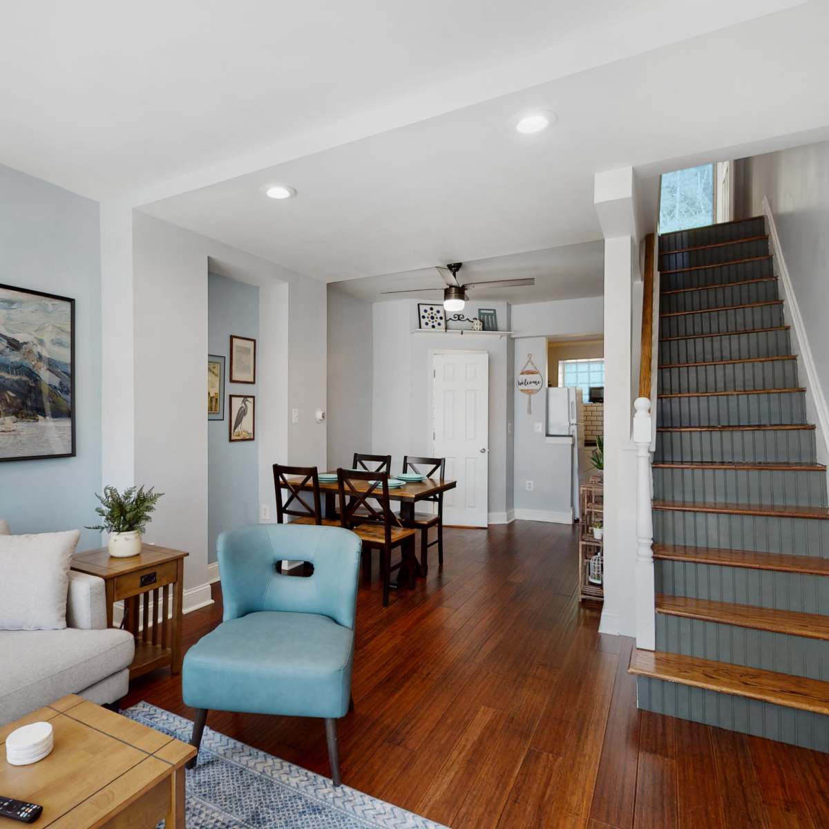 A modern living room with a couch, chair, and wooden table, leading to a dining area and a staircase.