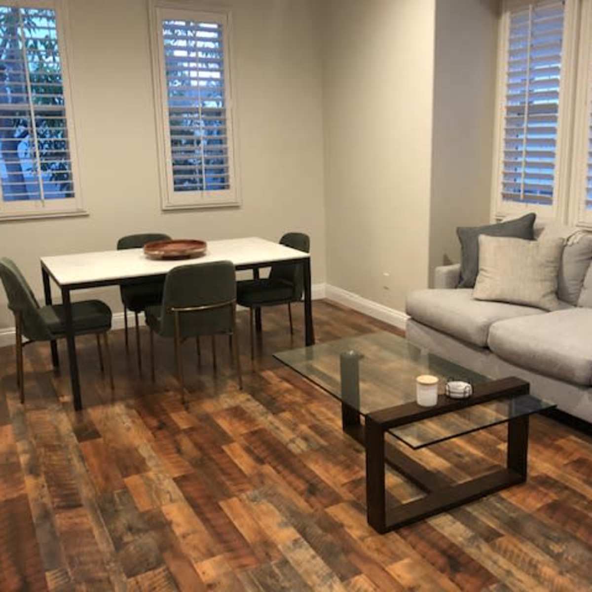 A room with a gray sofa, a glass coffee table, a white dining table surrounded by green chairs, and large windows with shutters.