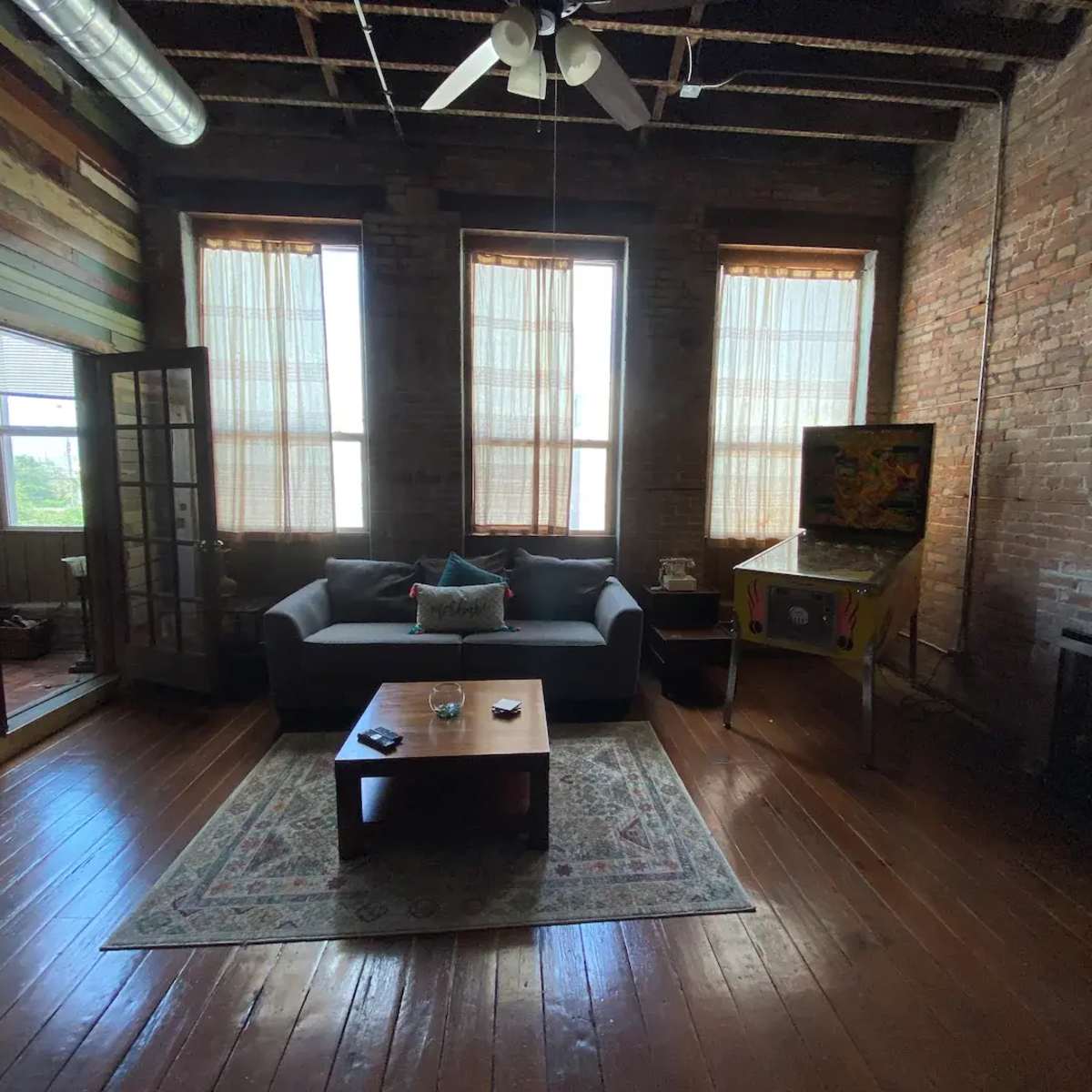 The room features a minimalist design with a sofa, a coffee table, a vintage pinball machine, and large windows displaying natural light, all set against exposed brick walls and wooden floors.