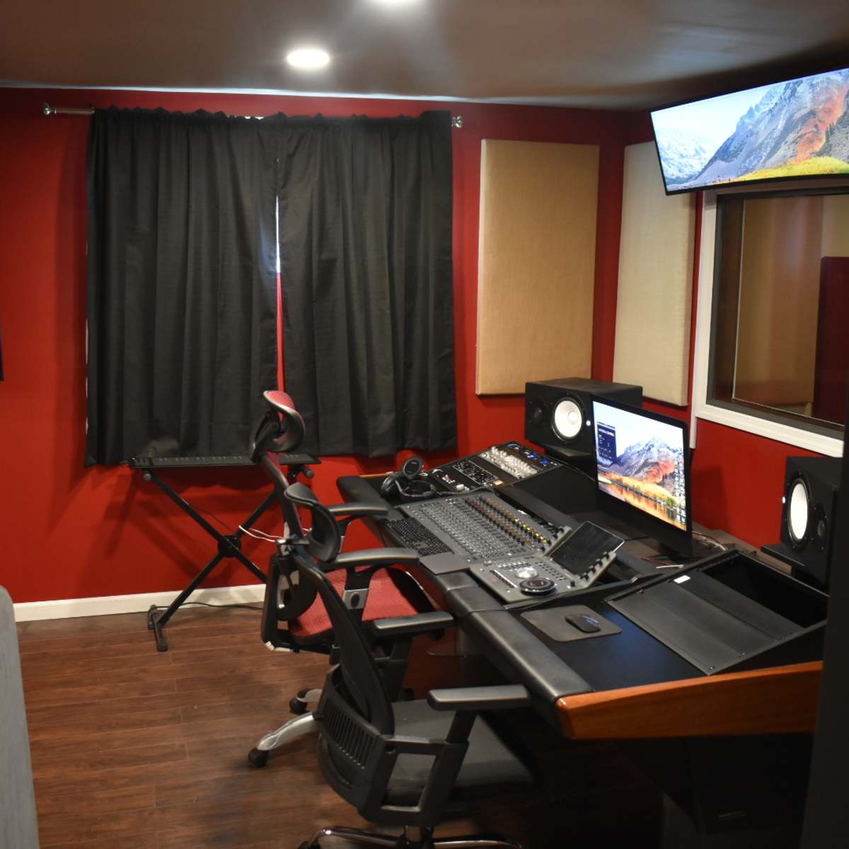 A small sound studio with a dark red wall, a control desk equipped with audio equipment, two chairs, and a large window, all illuminated by overhead lights.