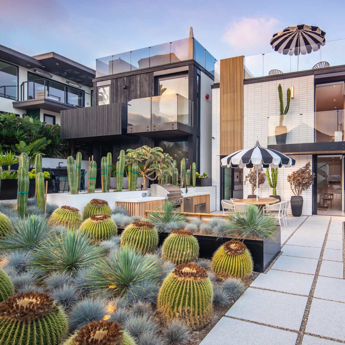 A modern house surrounded by landscaped gardens featuring cacti and an outdoor dining area with striped umbrellas.