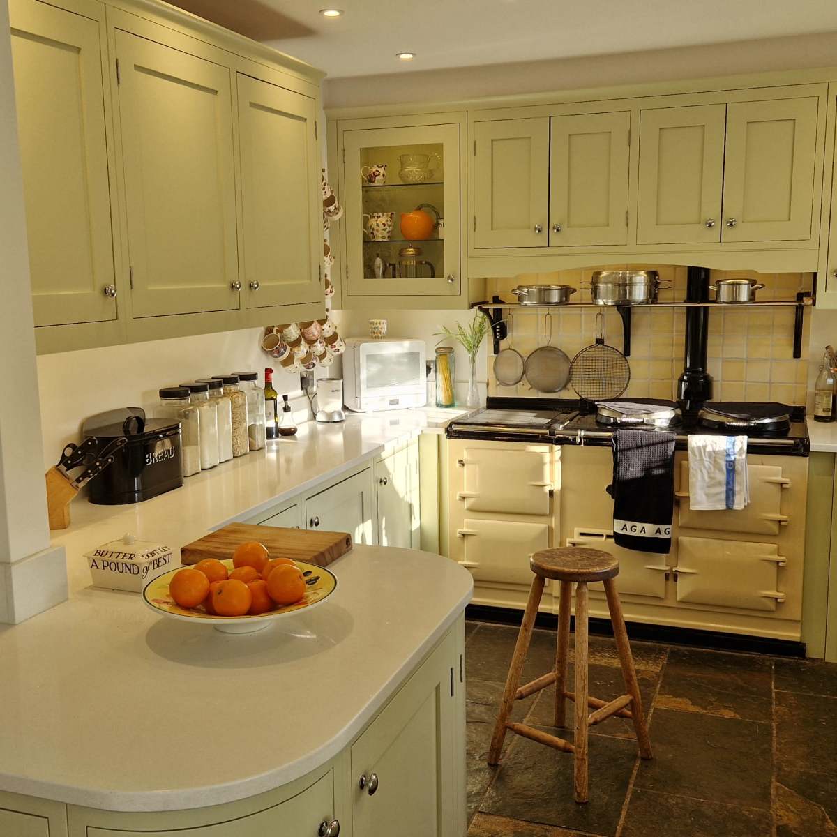 A brightly lit kitchen features green cabinets, a white countertop, an gas stove, and a round wooden stool beside a bowl of oranges.