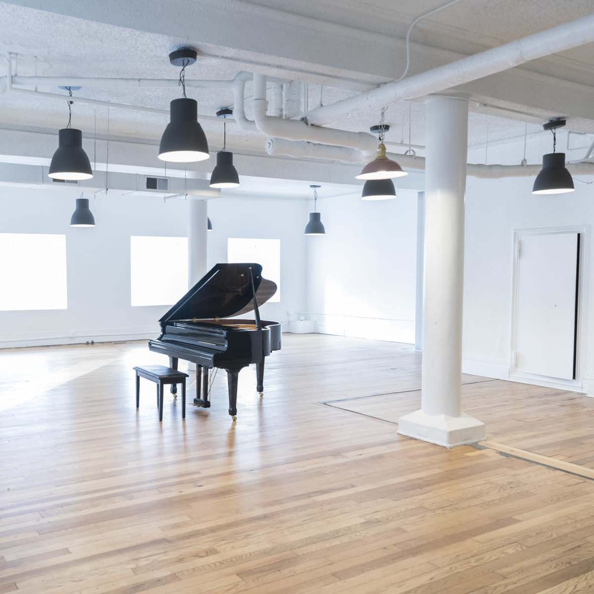 A grand piano sits alone in a spacious, brightly lit room with wooden floors and large windows.