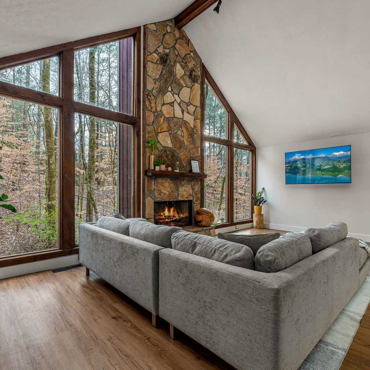 A modern living room features a stone fireplace and large windows overlooking a wooded area.