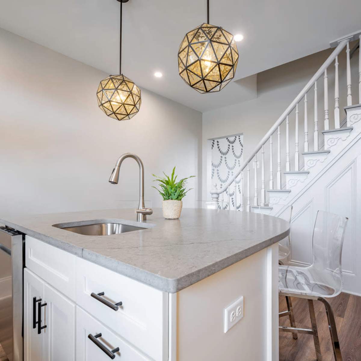 A modern kitchen area with a concrete countertop, a stainless steel sink, and two geometric pendant lights, adjacent to a staircase.