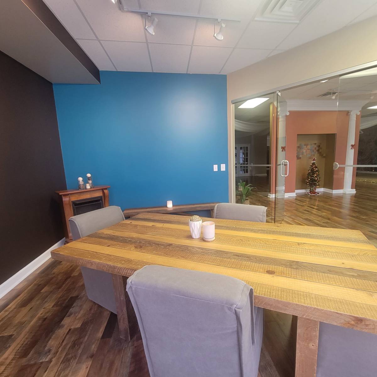 A wooden dining table with gray chairs is positioned in a room featuring blue and black accent walls, with a fireplace and a decorated Christmas tree visible in the background.