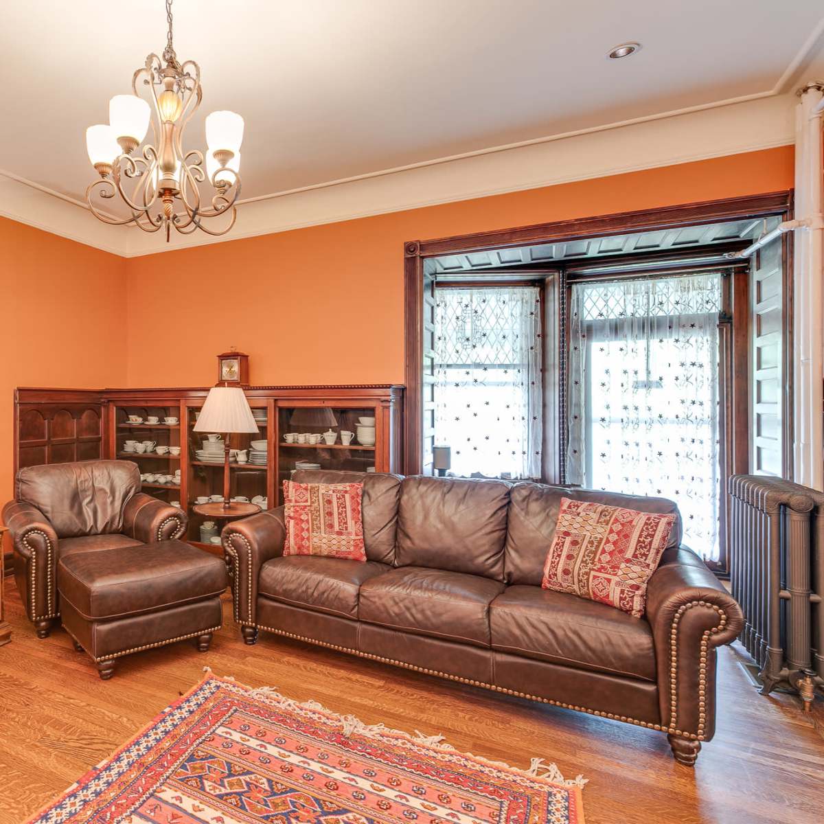 A warmly lit living room featuring brown leather sofas, a decorative mirror, a wooden cabinet, and a vibrant area rug on the hardwood floor.