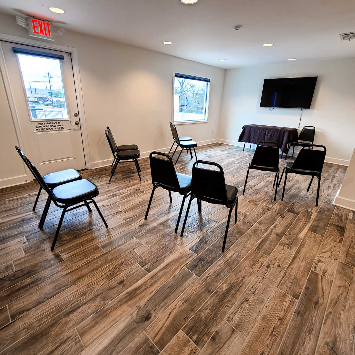 The room features six black chairs arranged around a table, with a television mounted on the wall and windows letting in natural light.