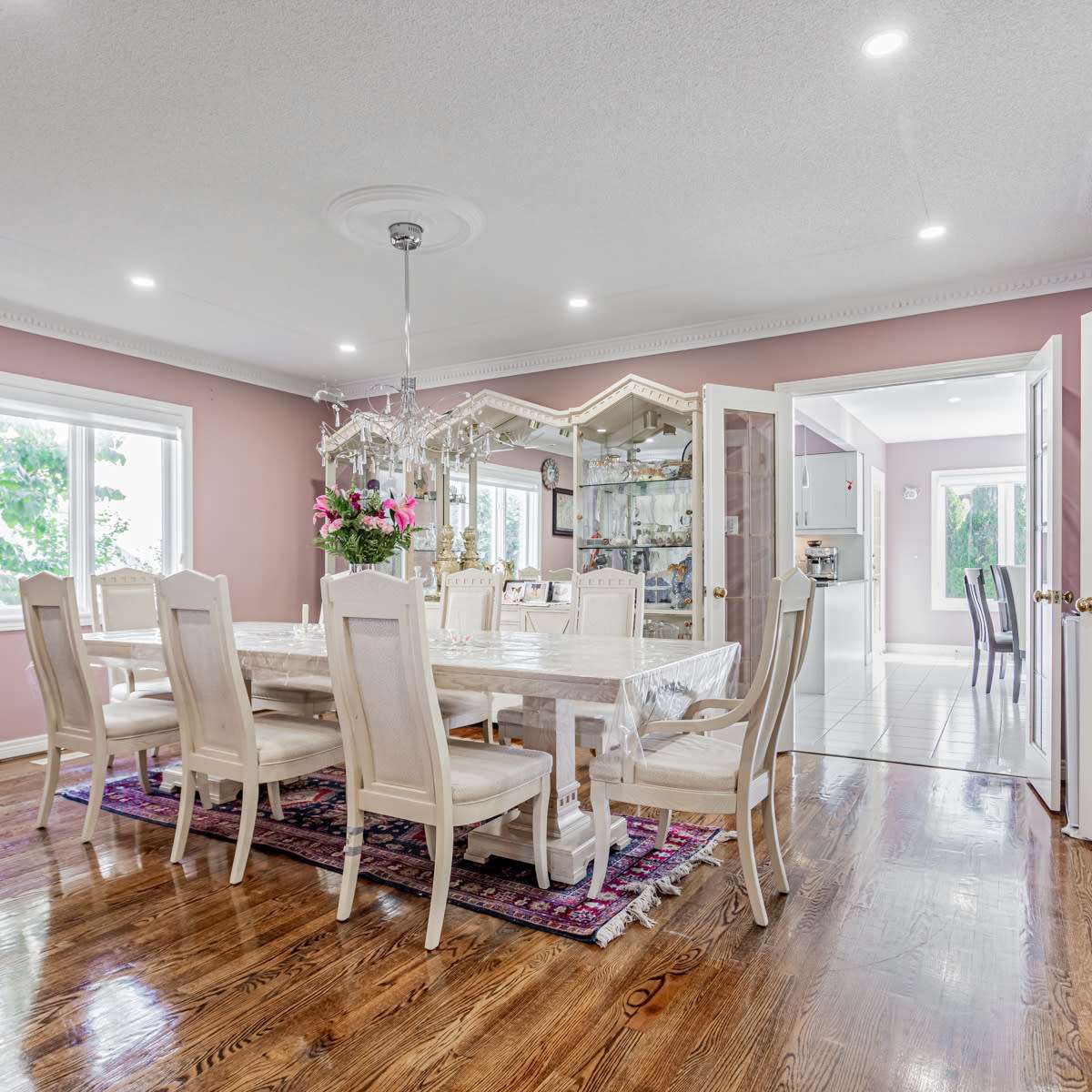 A spacious dining room features a large table surrounded by upholstered chairs, with a chandelier overhead and windows letting in natural light.