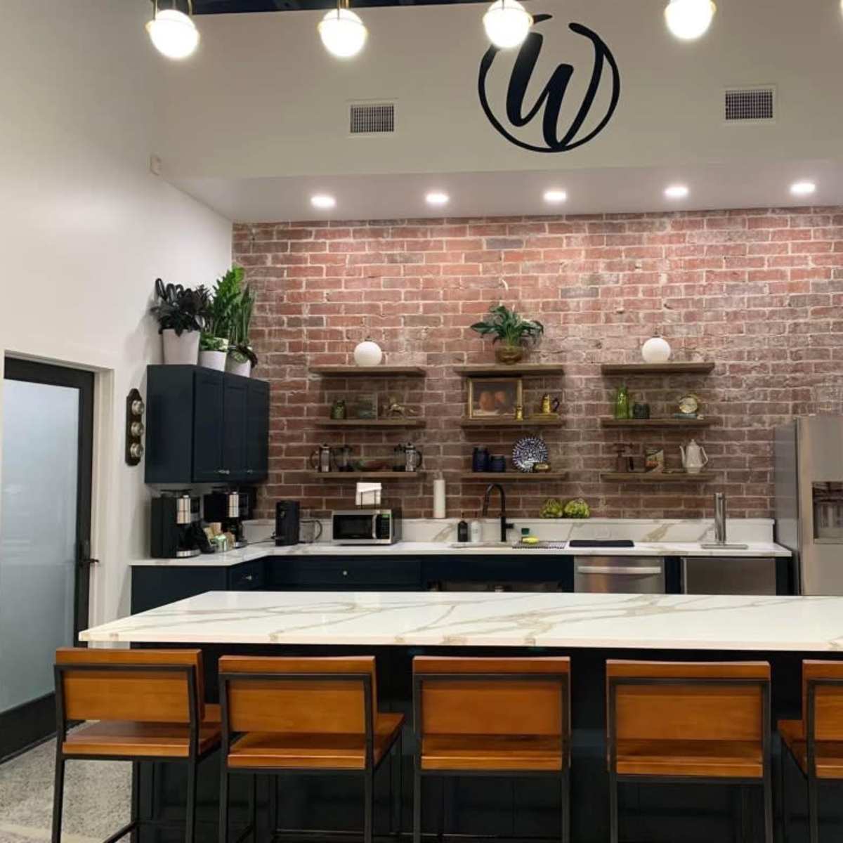 A modern kitchen with a brick accent wall, dark cabinetry, open shelves, and a large island with wooden bar stools.