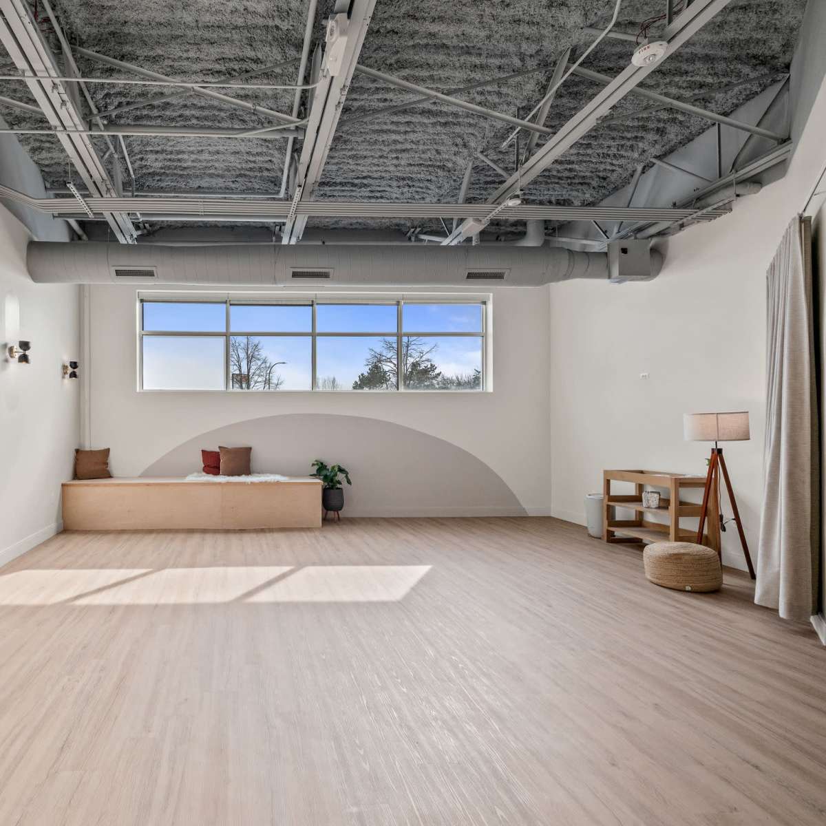 A spacious, well-lit room features a large window, wooden bench, and minimal furniture against a light-colored wall.