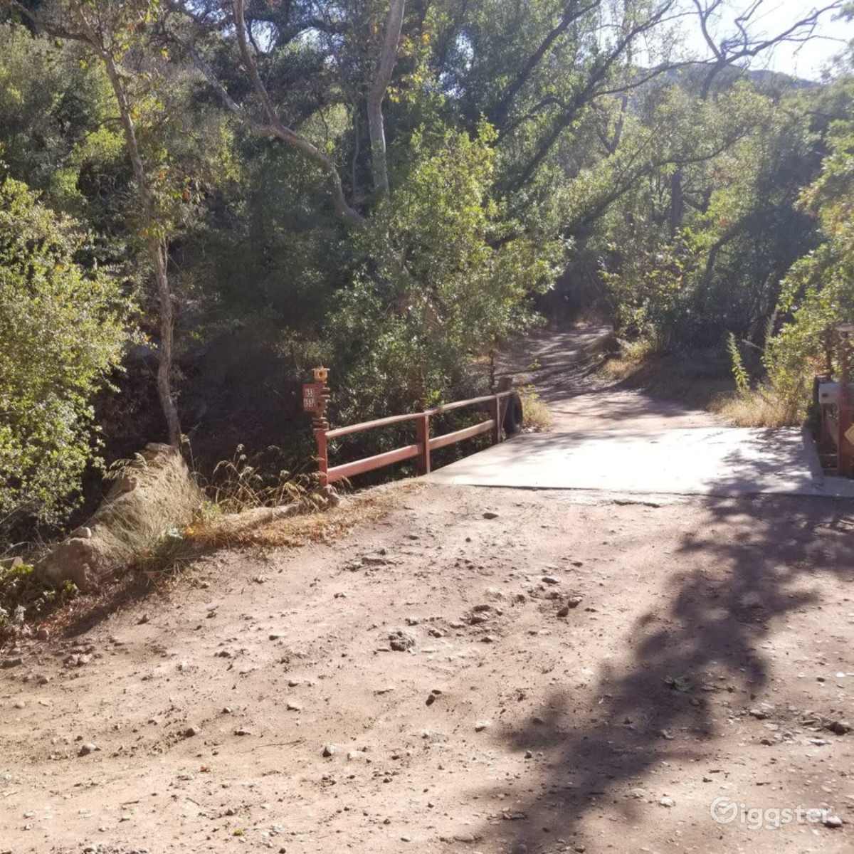 A wooden bridge leads over a dirt path surrounded by trees and sparse vegetation.