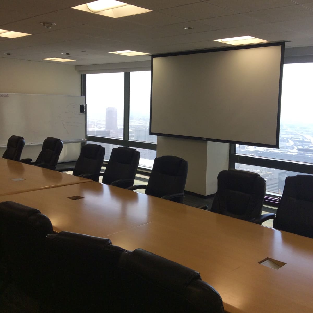 A conference room with a long wooden table and several black office chairs, featuring a large projector screen and windows overlooking a cityscape.