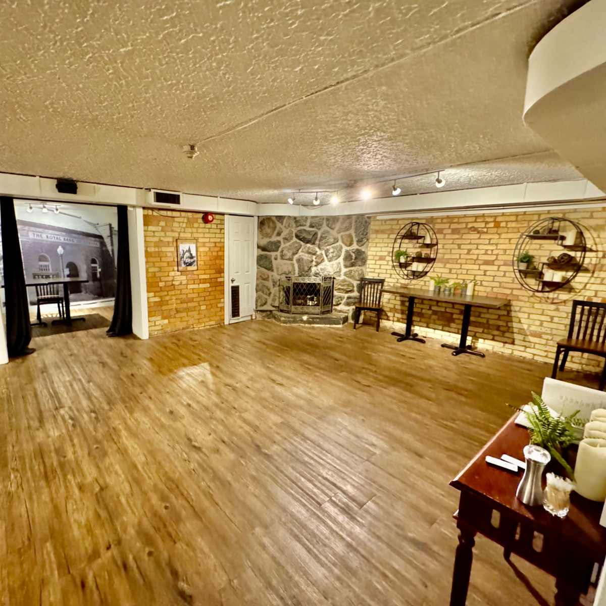 A spacious, empty room with wooden flooring, exposed brick walls, and a stone fireplace, featuring tables and chairs set against one wall.