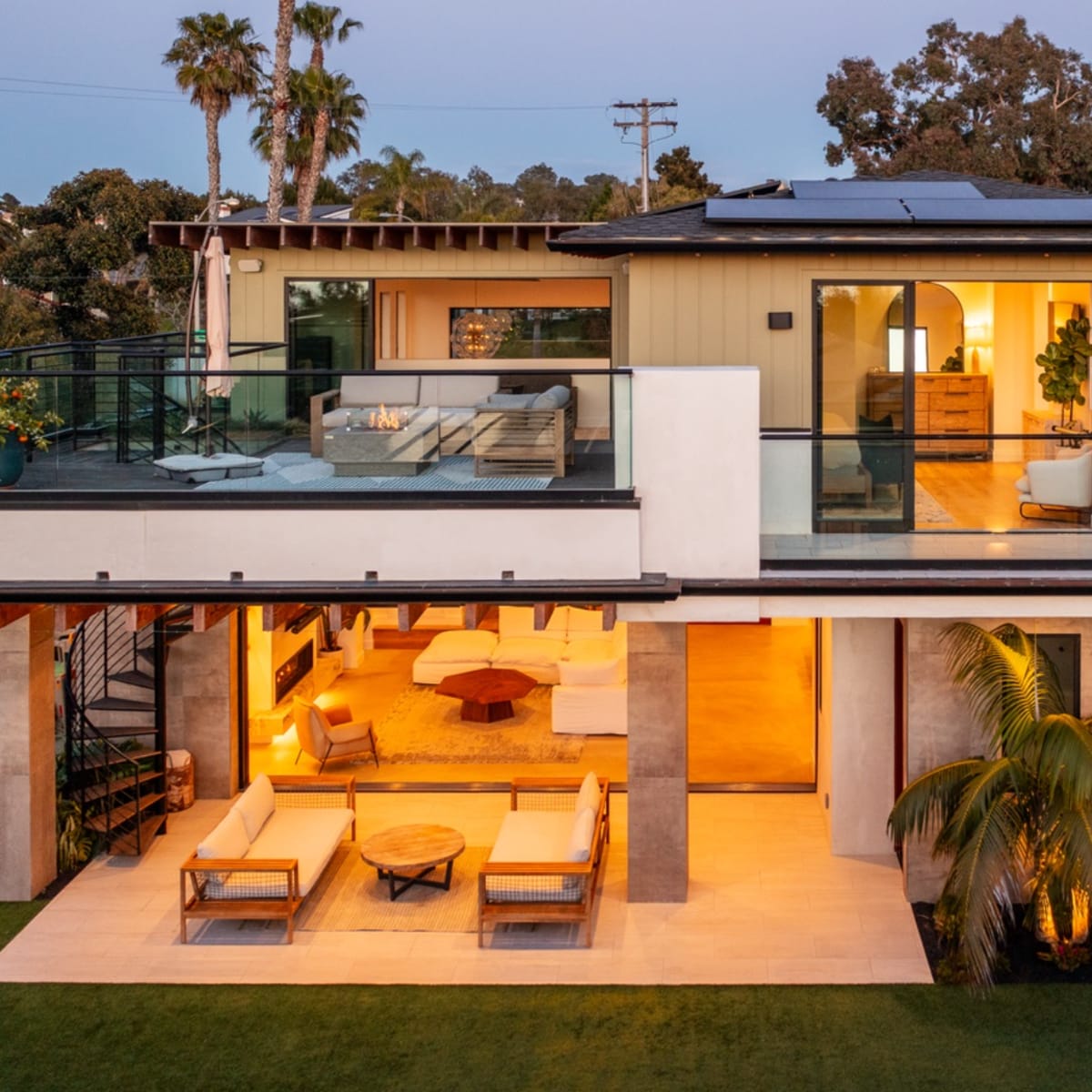 A modern two-story house features large glass windows, a spacious balcony with seating, and a well-lit interior visible from the patio.