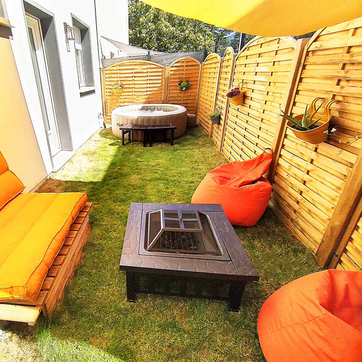 A small outdoor space with orange cushioned seating, a fire pit table, and scattered bean bags, surrounded by wooden fences and grass.