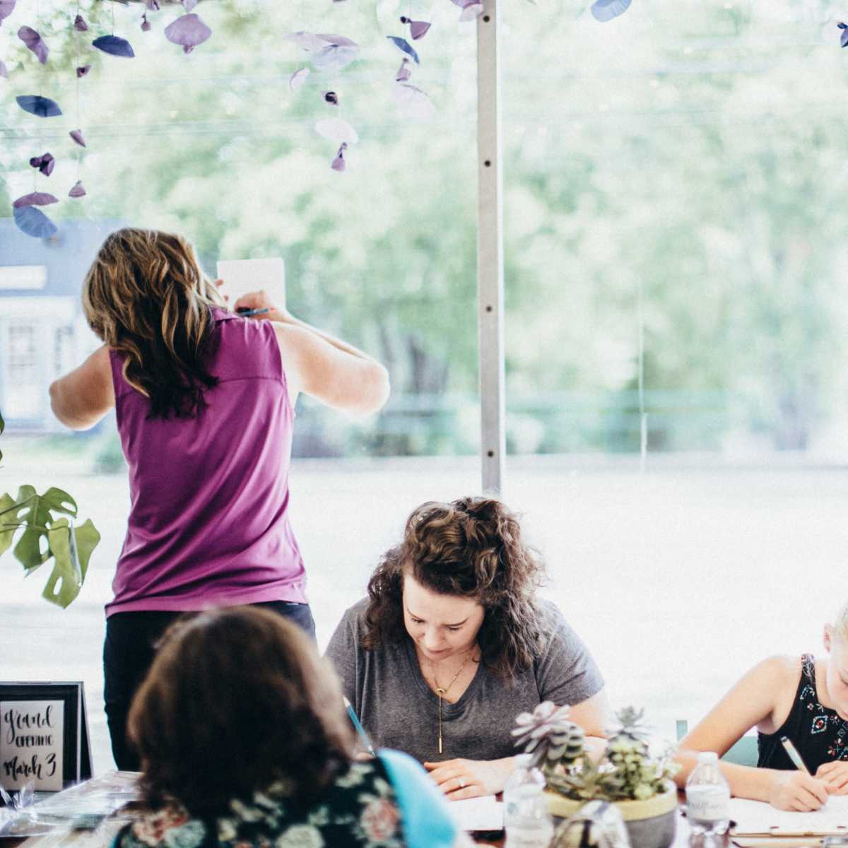 A group of people is engaged in writing and crafting activities at a table, while another person is hanging a paper on a wall.