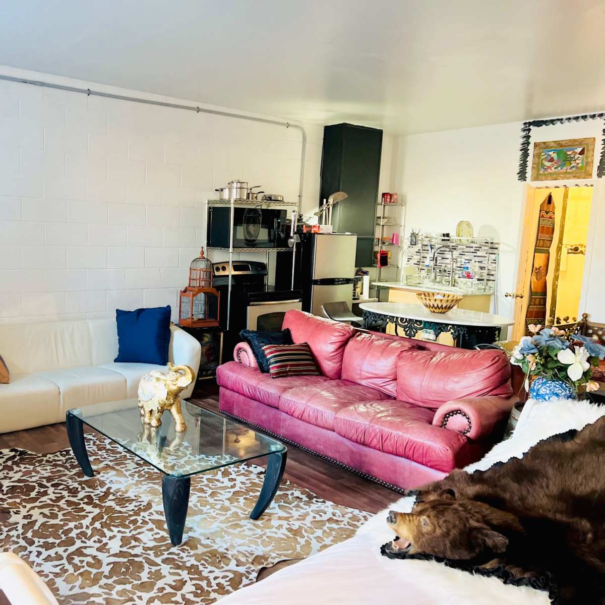 A cozy living room with a white couch, a pink leather sofa, a glass coffee table on a patterned rug, and a view of a kitchen area in the background.