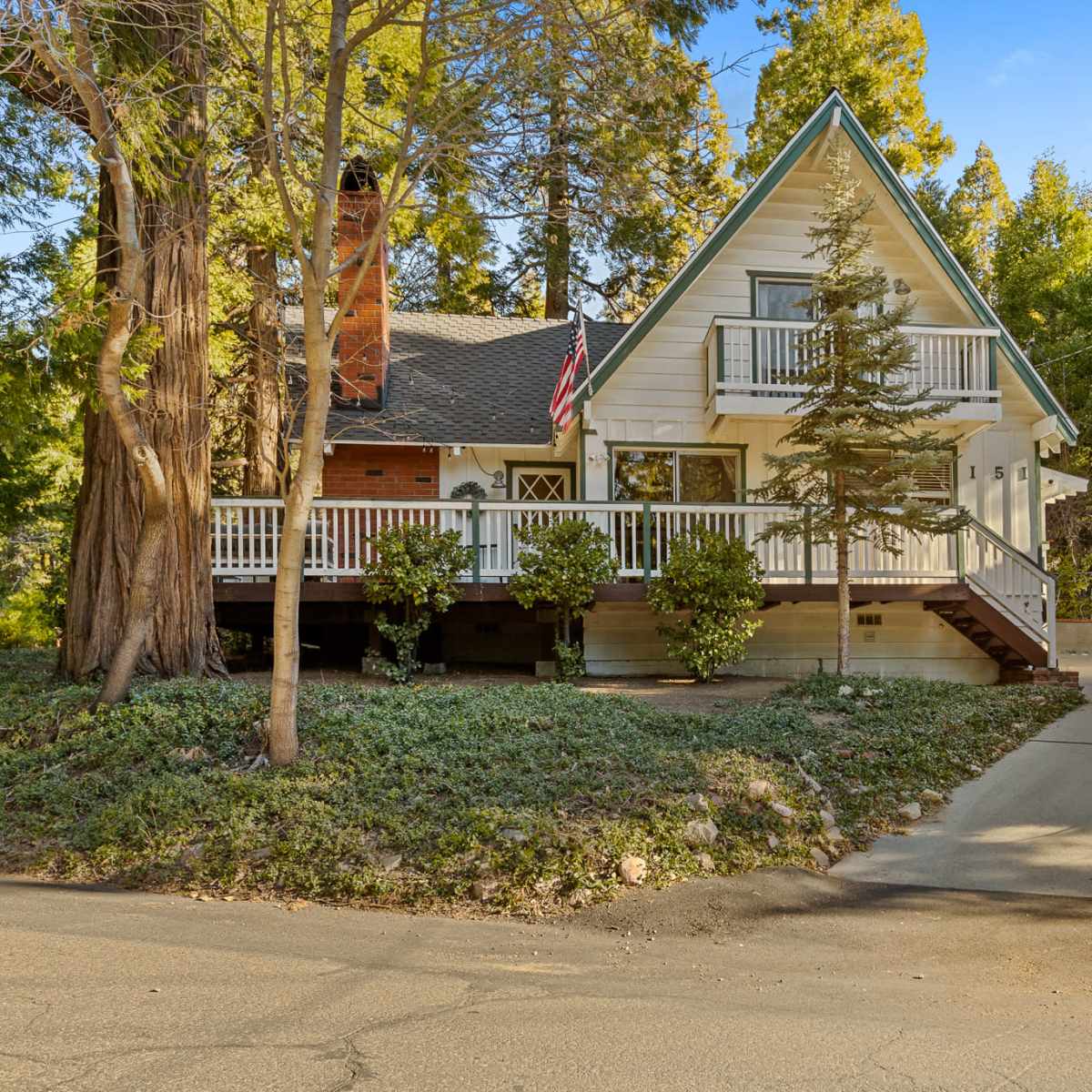 A two-story house with a peaked roof is set among trees, featuring a front porch and a sloped driveway leading to the street.