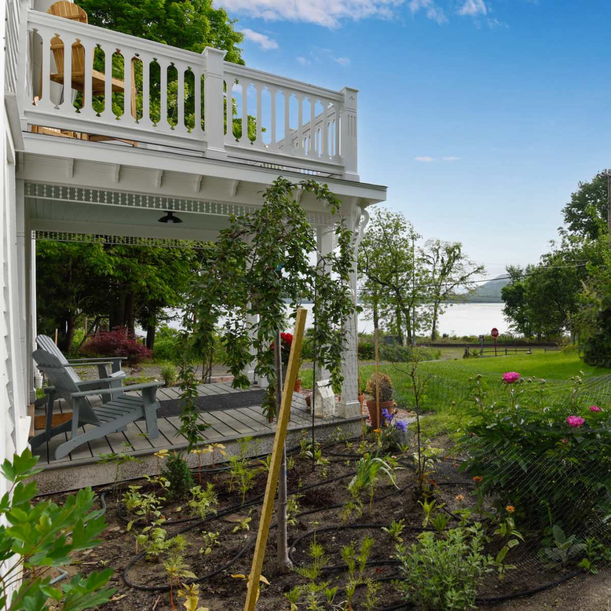 A house with a porch and a garden overlooks a lake, surrounded by trees and a grassy yard.
