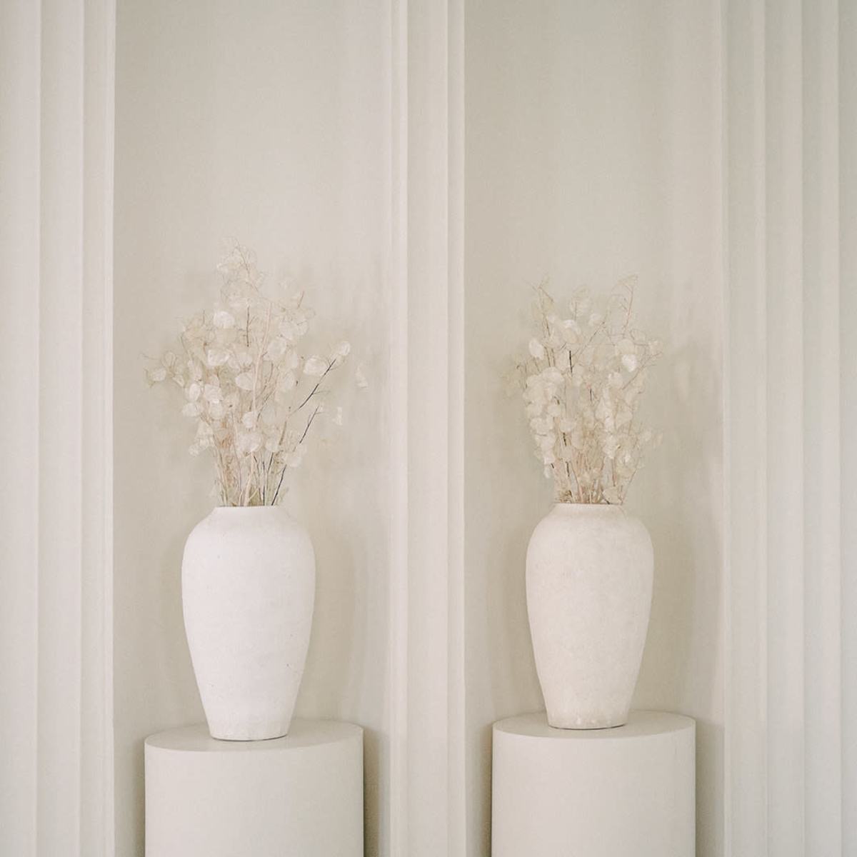 Two white vases filled with dried flowers are placed on cylindrical pedestals against a backdrop of vertical white panels.